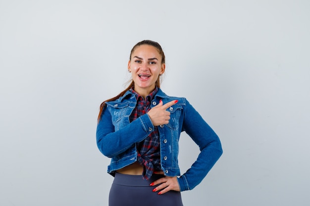 Jovem mulher com camisa quadriculada, jaqueta, calça apontando para o lado direito, mantendo a mão no quadril e olhando espantada, vista frontal.