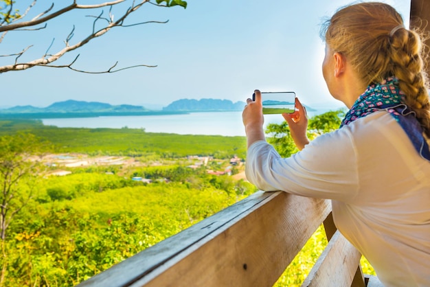 Jovem mulher com celular tirando imagem da paisagem marítima da ilha montanhosa da Tailândia