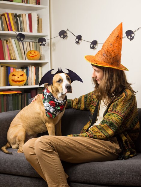 Jovem mulher com chapéu de bruxa de halloween sente-se com seu cão de estimação vestido para festa em casa