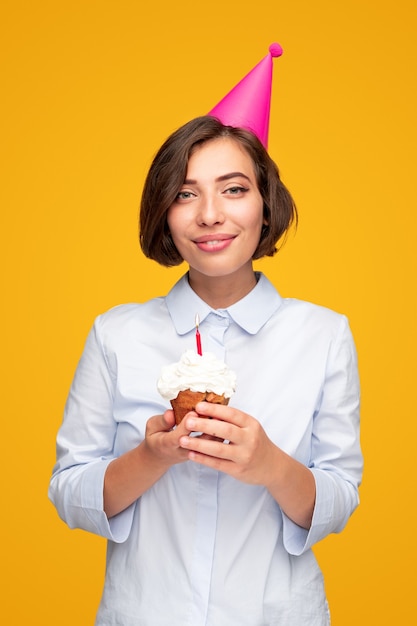 Jovem mulher com chapéu de festa sorrindo e olhando para a câmera enquanto saboreia bolinho com vela durante a festa de aniversário contra um fundo amarelo