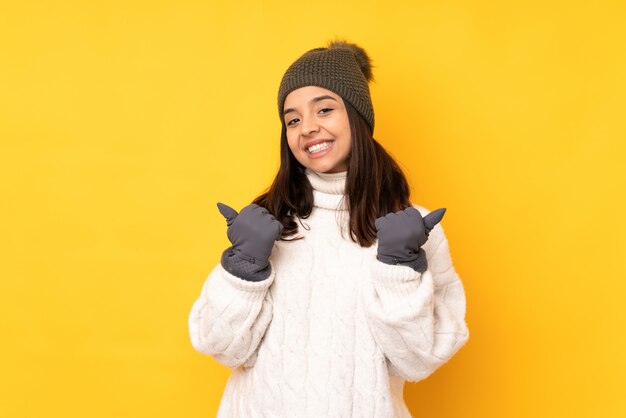 Jovem mulher com chapéu de inverno sobre parede amarela isolada com polegares para cima gesto e sorrindo