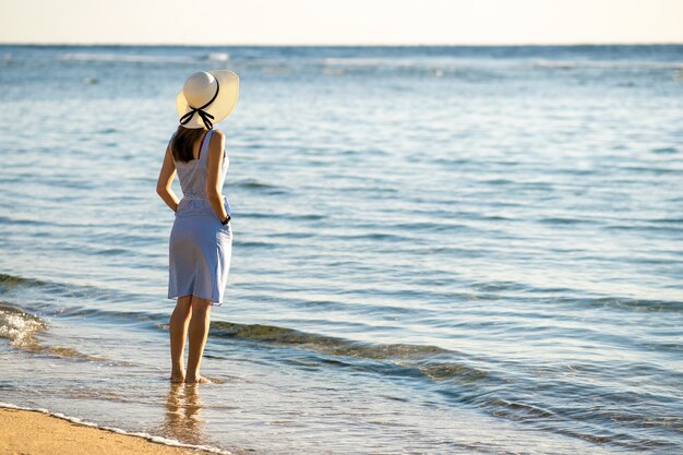 Jovem mulher com chapéu de palha e um vestido sozinho na praia de areia vazia na costa do mar.