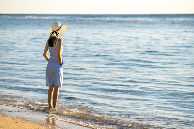 Jovem mulher com chapéu de palha e um vestido sozinho na praia de areia vazia na costa do mar.