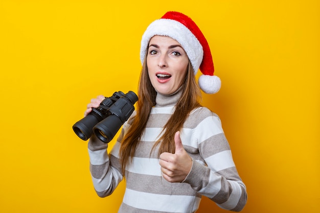 Jovem mulher com chapéu de Papai Noel com binóculos em um fundo amarelo.