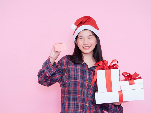 Jovem mulher com chapéu de papai noel segurando caixas de cartão e presente de crédito na parede rosa.