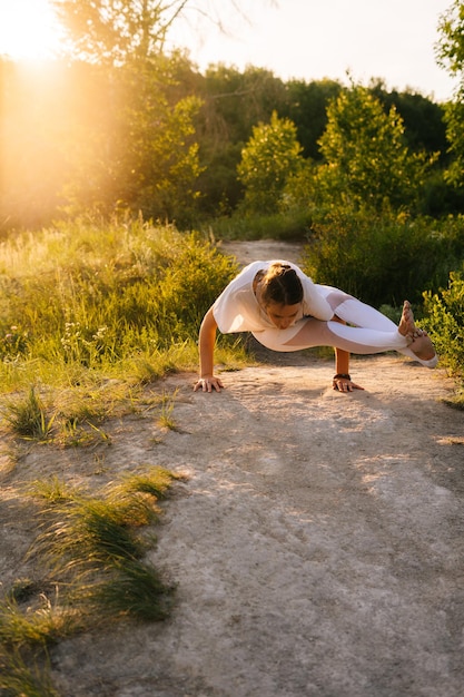 Jovem mulher com corpo alongado praticando ioga e realizando pose de oito ângulos fora no parque à noite no fundo da luz solar Mulher fazendo exercícios avançados de ioga ao ar livre na grama verde