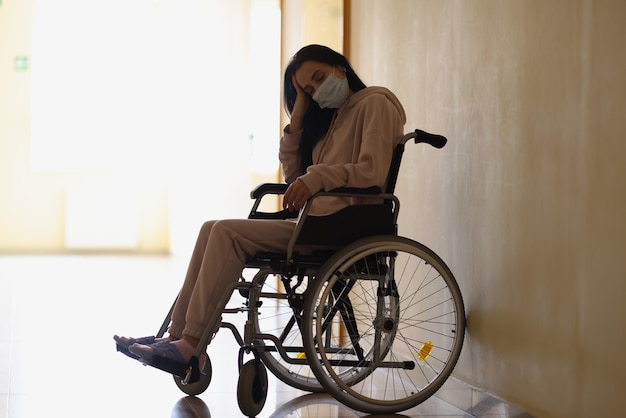 Jovem mulher com deficiência usando máscara protetora médica, sentada em uma cadeira de rodas no corredor do hospital