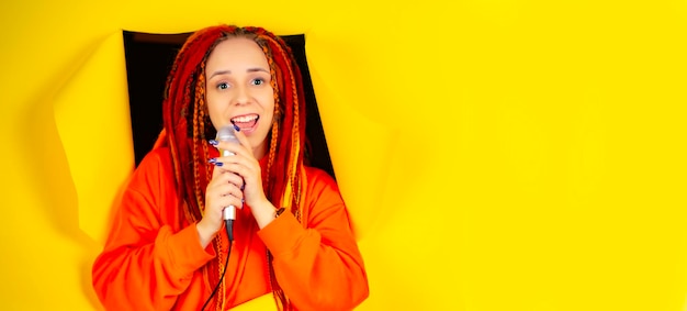 Jovem mulher com dreadlocks brilhantes cantando no microfone saindo do buraco no fundo amarelo Cantora emocional com microfone no estúdio com espaço para o seu texto