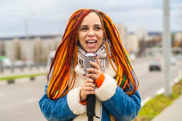 Jovem mulher com dreadlocks brilhantes com microfone retrô na rua da cidade Retrato de cantora cantando no microfone em tempo nublado