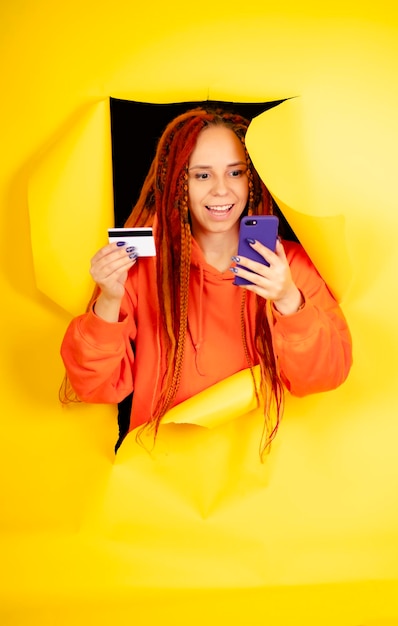 Jovem mulher com dreadlocks cartão de crédito do telefone móvel saindo do buraco no fundo amarelo Navegação feminina positiva smartphone inserindo detalhes do cartão pagando compra na internet
