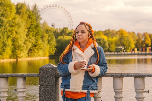 Jovem mulher com dreadlocks em roupas quentes em pé perto do lago tomando café no parque