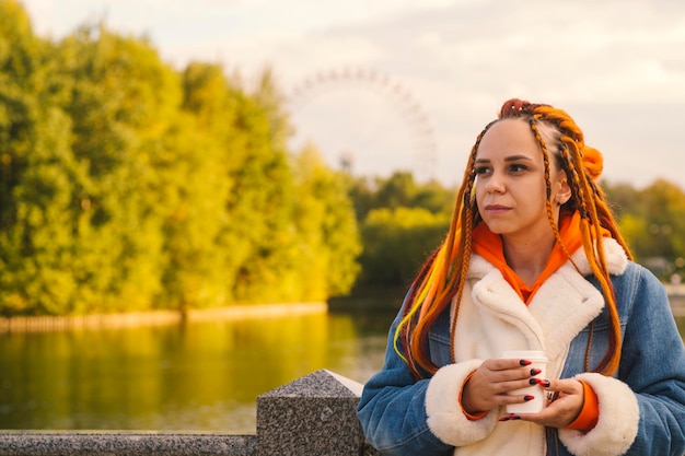 Jovem mulher com dreadlocks em roupas quentes em pé perto do lago tomando café no parque