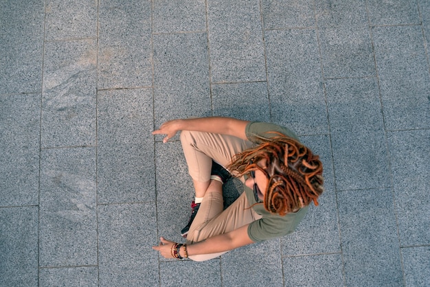 Foto jovem mulher com dreadlocks vermelhos se senta em um ladrilho cinza na rua e medita. atirando de cima. garota solitária