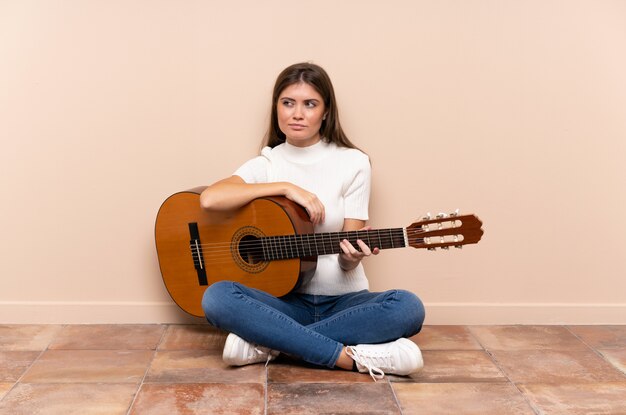 Jovem mulher com guitarra, sentada no chão em pé e olhando para o lado