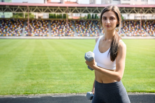 Jovem mulher com halteres posando do lado de fora. Ela está fazendo exercícios matinais ao ar livre