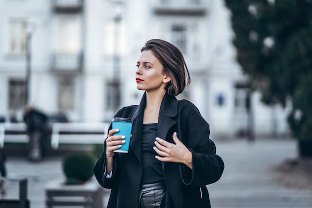 Jovem mulher com lábios vermelhos e cabelo curto, vestida de preto, posando, segurando uma xícara de café nas mãos. Por trás do fundo urbano turva.