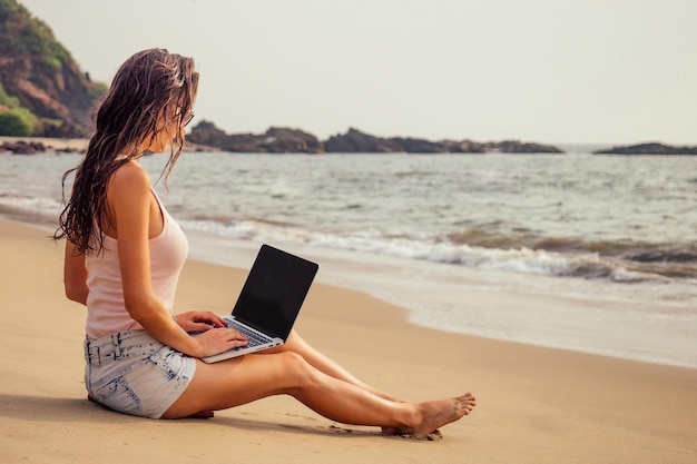 Jovem mulher com laptop de férias tropicais, sentado na areia em uma ilha deserta e paradisíaca na praia à beira-mar. Surfista freelancer de menina no Oceano Índico. Trabalho remoto e freelancer de liberdade.