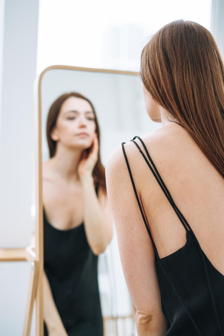Jovem mulher com longos cabelos escuros na noite elegante vestido preto com costas abertas perto do espelho no interior branco pessoas por trás