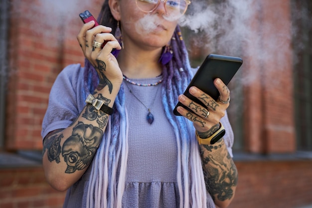 Foto jovem mulher com longos dreadlocks enviando mensagens de texto em smartphone e fumando