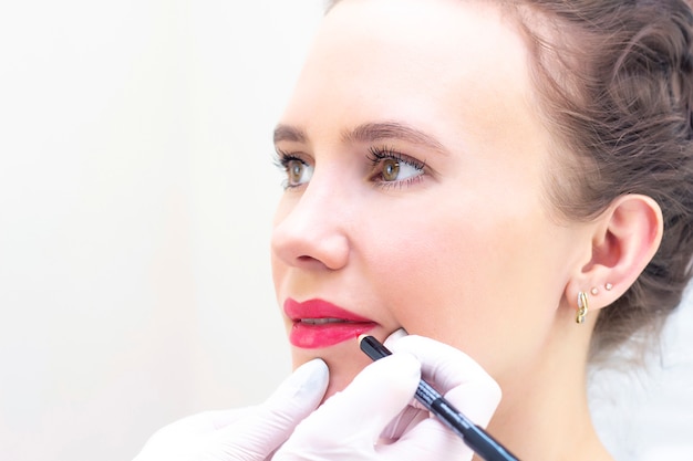 Foto jovem mulher com maquiagem permanente nos lábios no salão de esteticistas. maquiagem permanente (tatuagem). desenhando um contorno com um lápis labial branco