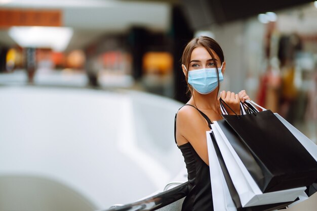 Jovem mulher com máscara médica protetora estéril no rosto com sacolas de compras no shopping.