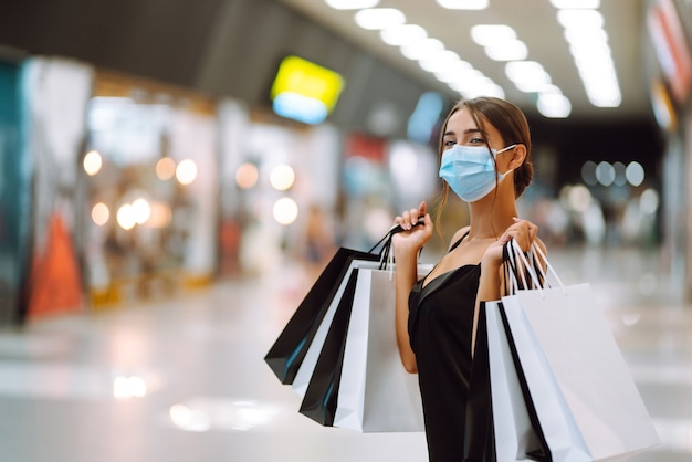 Jovem mulher com máscara médica protetora estéril no rosto com sacolas de compras no shopping.