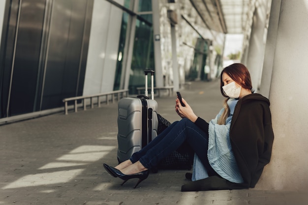 Jovem mulher com máscara médica senta-se perto de bagagem no aeroporto. Mulher esperando o voo, usando um telefone celular e olhando para a câmera.