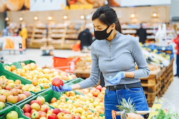 Jovem mulher com máscara protetora e luvas, escolhendo frutas em um supermercado ou mercado.