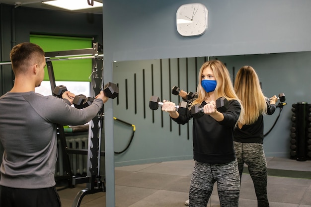 Jovem mulher com máscara protetora malhando com o personal trainer na academia durante a pandemia de Covid-19. Ela está bombeando seus músculos com halteres. Foco suave