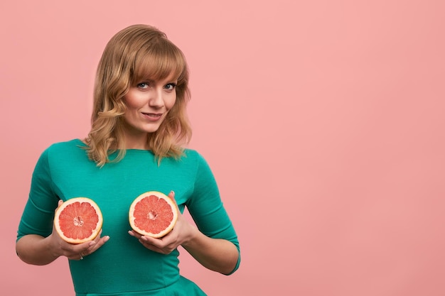 Jovem mulher com metades de toranja nas mãos menina alegre em verde posando para a câmera com toranjas em um espaço de cópia de fundo rosa