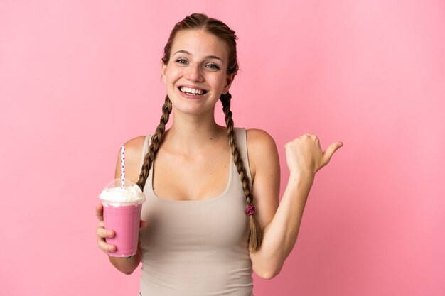 Jovem mulher com milk-shake de morango isolado em fundo rosa apontando para o lado para apresentar um produto