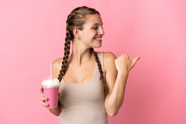 Jovem mulher com milkshake de morango isolado em rosa apontando para o lado para apresentar um produto