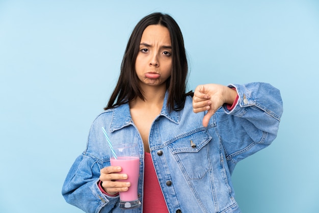 Jovem mulher com milkshake de morango no azul isolado, fazendo bom-mau sinal. Indeciso entre sim ou não