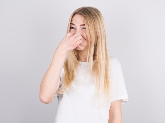 Jovem mulher com nojo no rosto aperta o nariz na parede branca. Expressão facial da emoção negativa.