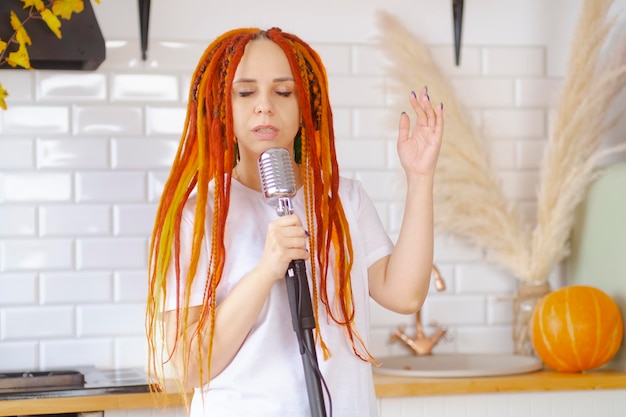 Jovem mulher com penteado brilhante com microfone retrô na cozinha Retrato de cantora com dreadlocks cantando no microfone em casa