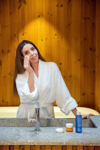 Jovem mulher com roupão de banho branco, enxugando a pele do rosto com algodão e produto cosmético durante procedimentos diários de beleza no banheiro
