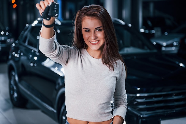 Jovem mulher com roupas casuais em pé perto de um carro preto moderno com as chaves nas mãos.