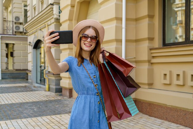 Jovem mulher com sacolas de compras fazendo selfie andando por uma cidade em dia de verão