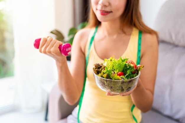 Jovem mulher com salada caseira saudável, exercitando-se com halteres em casa.