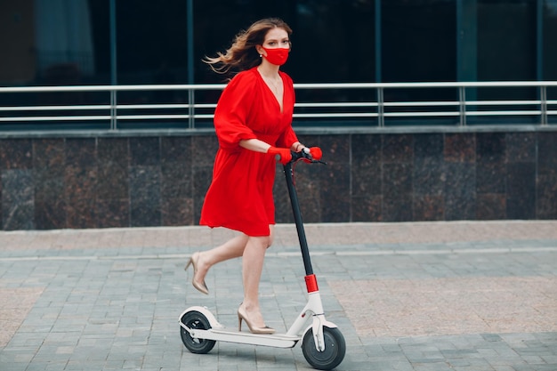 Jovem mulher com scooter elétrico de vestido vermelho e luvas com máscara facial na cidade Nova moda normal e conceito de proteção COVID coronavírus