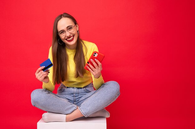 Jovem mulher com telefone e cartão de banco em fundo vermelho