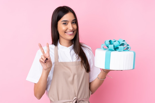 Jovem mulher com um bolo grande sobre parede isolada, sorrindo e mostrando sinal de vitória