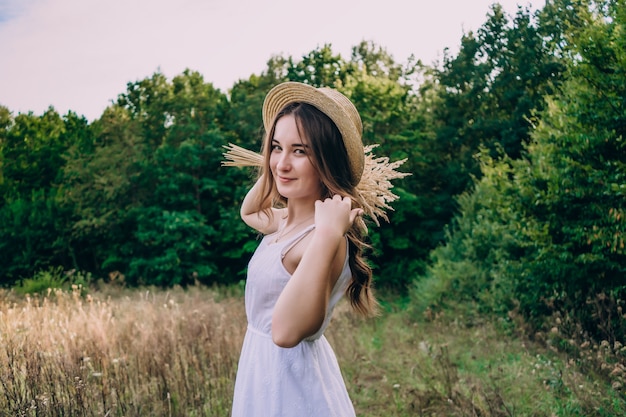 Jovem mulher com um chapéu de palha em um fundo de ervas altas. linda garota com um vestido com um buquê de flores secas no campo.