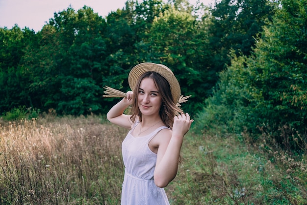 Jovem mulher com um chapéu de palha em um fundo de ervas altas. Linda garota com um vestido com um buquê de flores secas no campo.