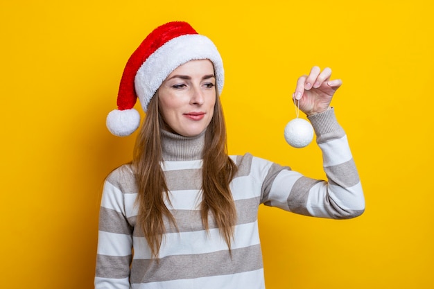 Jovem mulher com um chapéu de Papai Noel segura uma bola decorativa em um fundo amarelo.