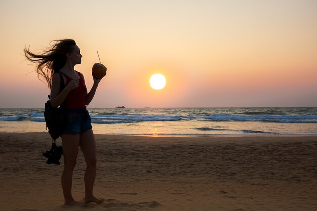 Jovem mulher com um coco olhando o pôr do sol