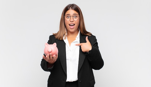 Jovem mulher com um cofrinho se sentindo feliz, surpresa e orgulhosa, apontando para si mesma com um olhar animado e surpreso