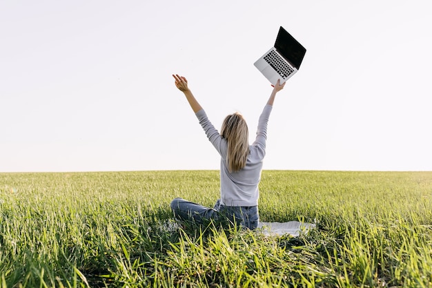 Jovem mulher com um laptop, sentada em campo com grama verde fresca.