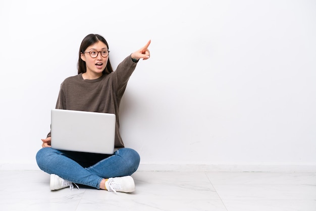 Jovem mulher com um laptop sentada no chão apontando para longe