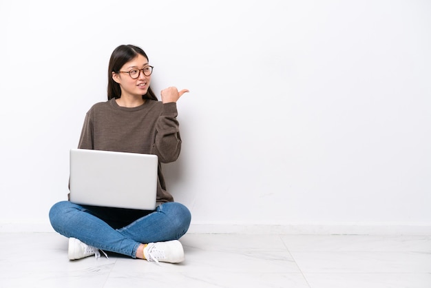 Jovem mulher com um laptop sentado no chão, apontando para o lado para apresentar um produto